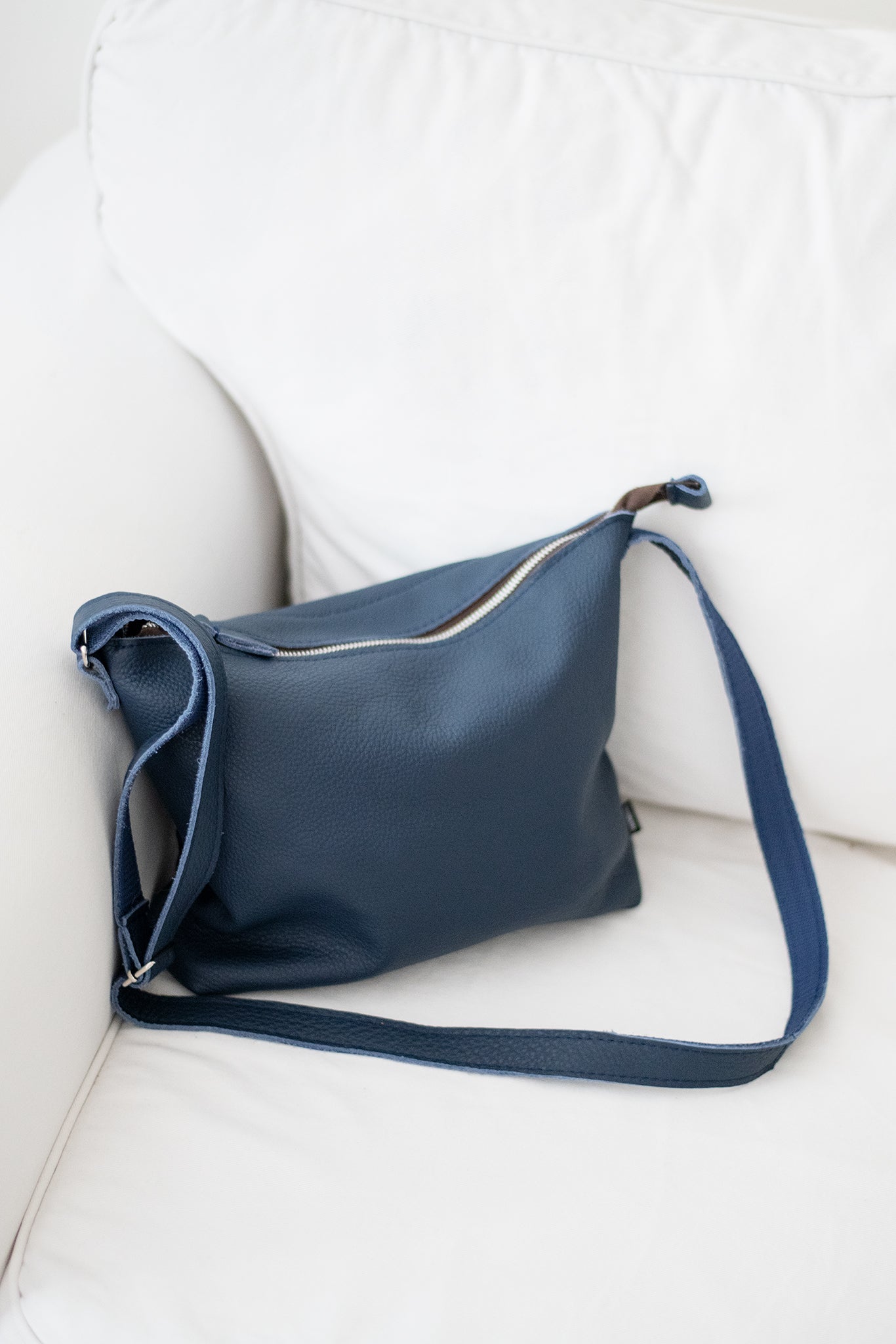 Blue leather shoulder bag with adjustable strap placed on white couch by Trendbag