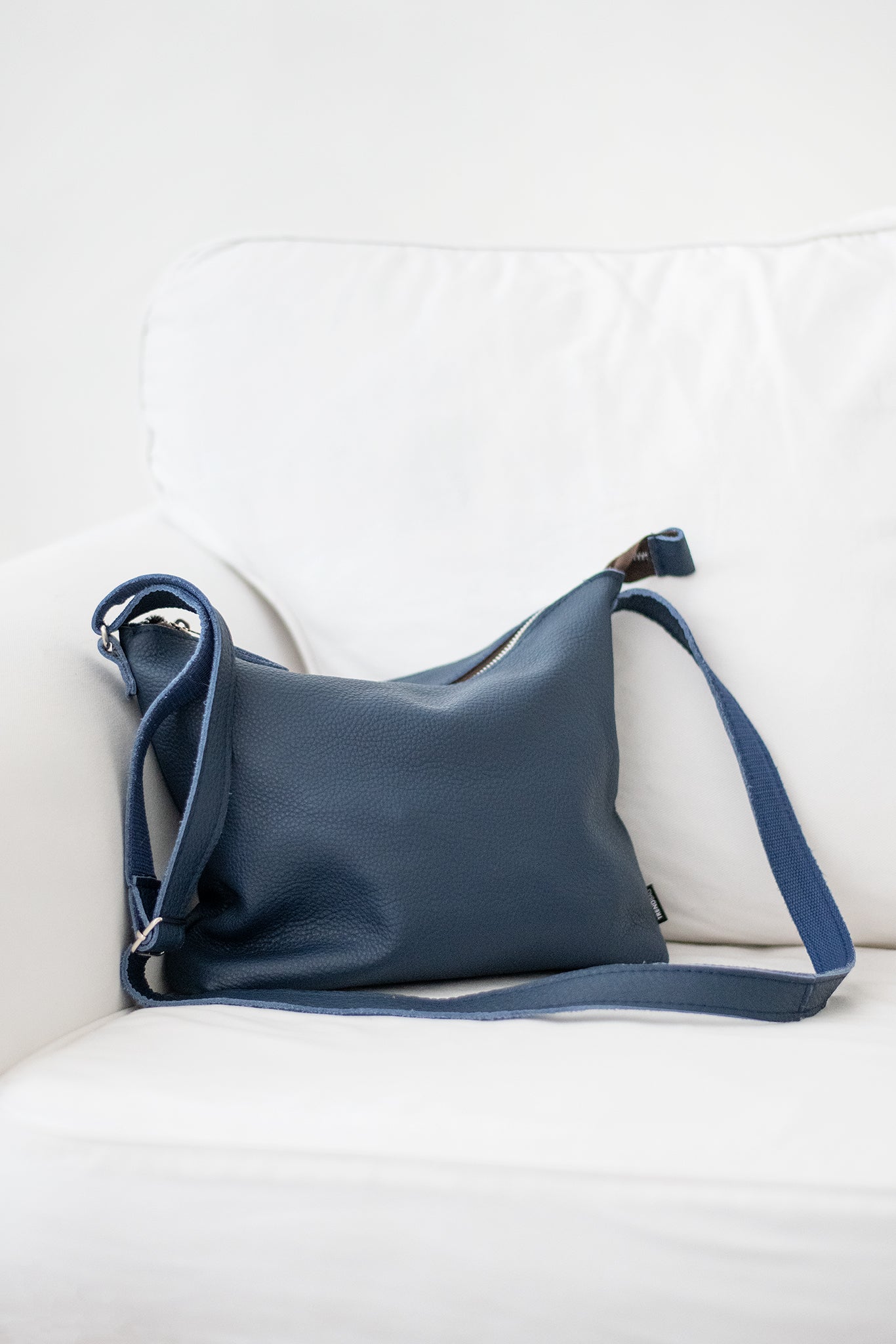 Dark blue leather shoulder bag by Trendbag resting on a white couch with adjustable strap